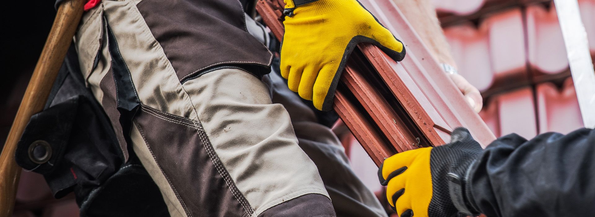 Zwei Personen mit gelben Handschuhen heben gemeinsam mehrere lange Holzbretter
